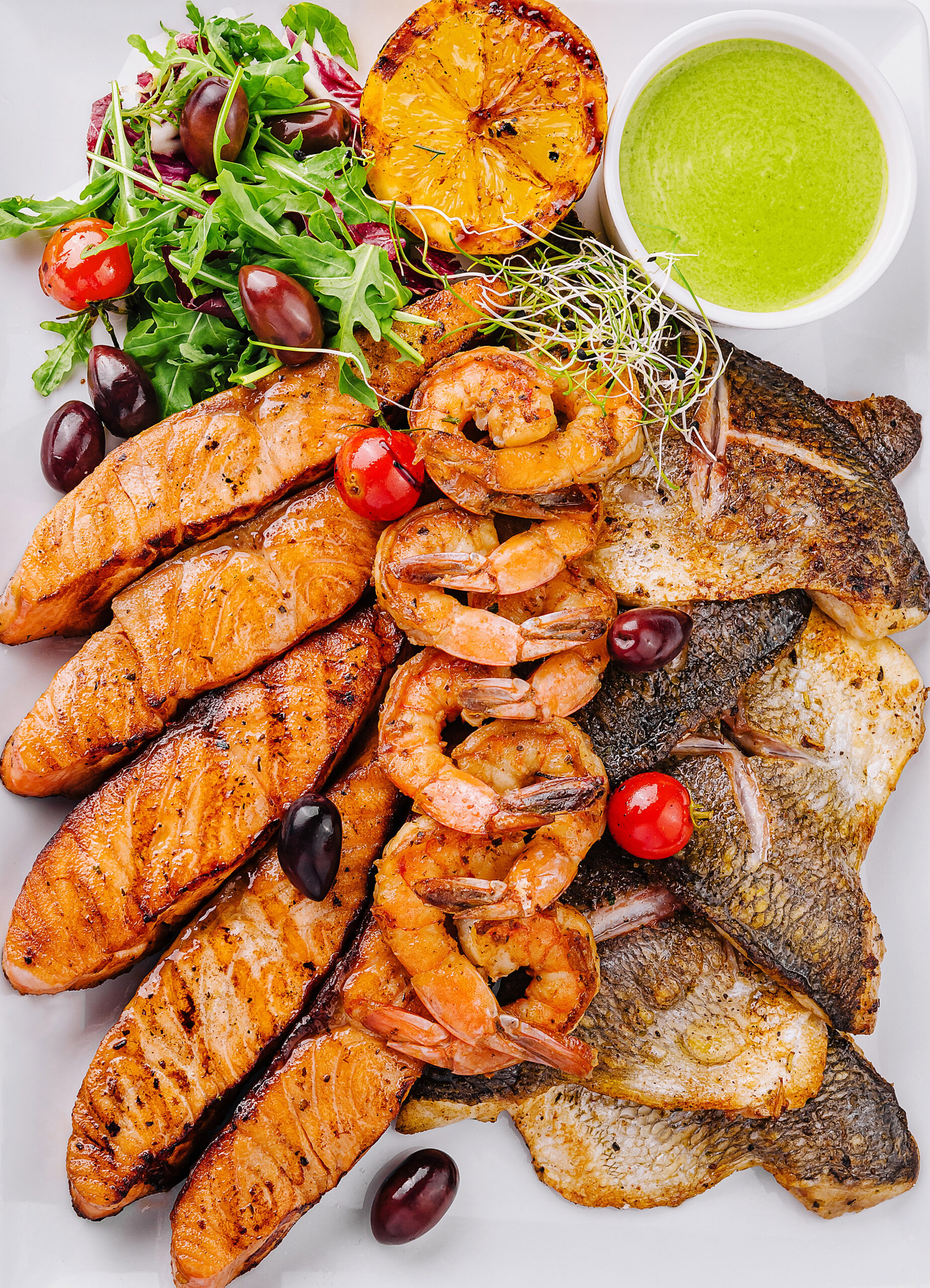 Gourmet seafood platter featuring grilled fish, shrimp, and salad served with sauce on a white background
